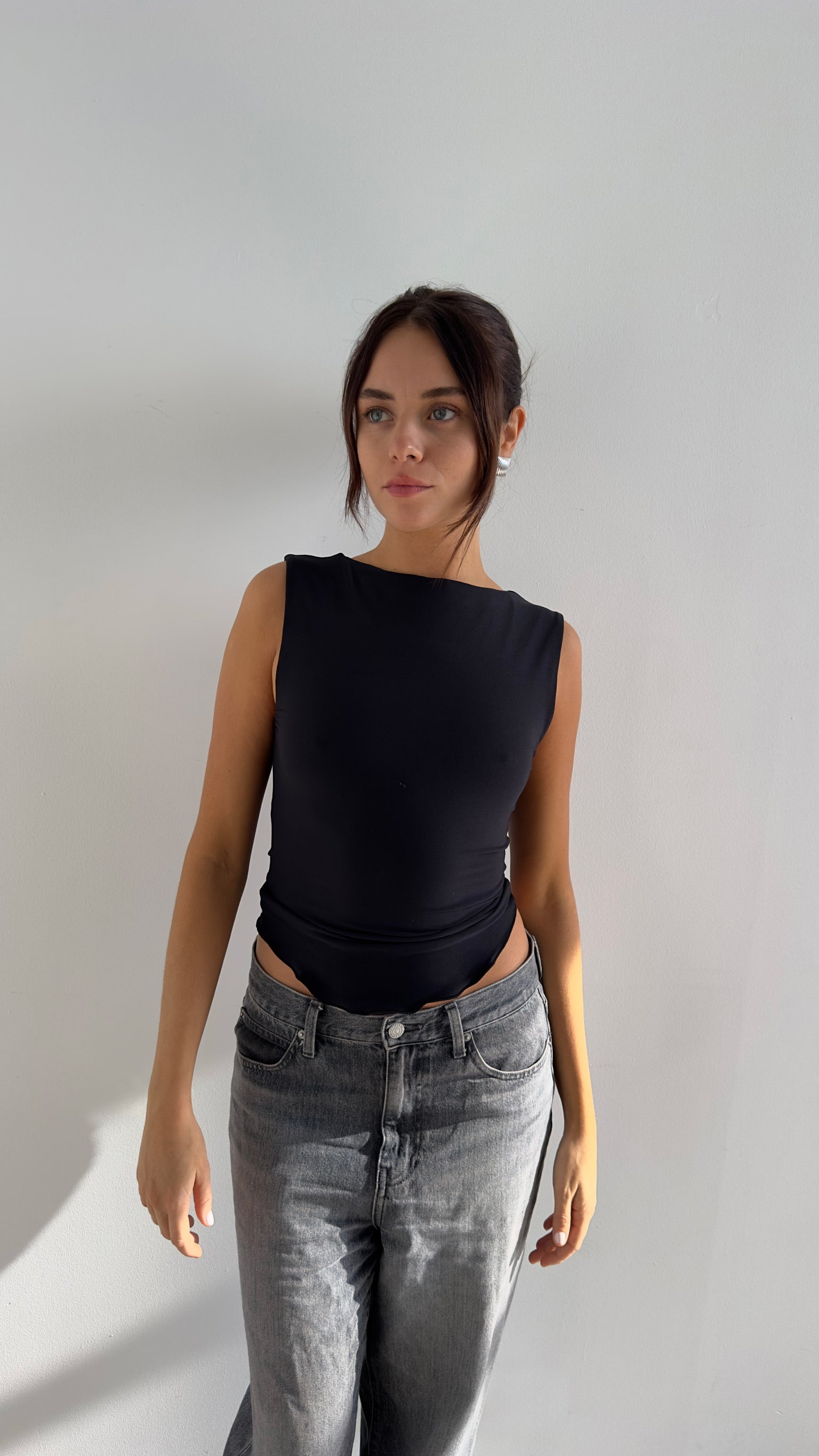 A female model wearing a black, sleeveless top with a boat neckline and fitted waist, made from a stretchy, recycled fabric, standing against a neutral background.