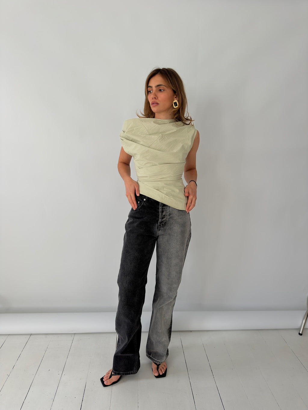 Woman wearing a light green sleeveless top and dark jeans against a white background